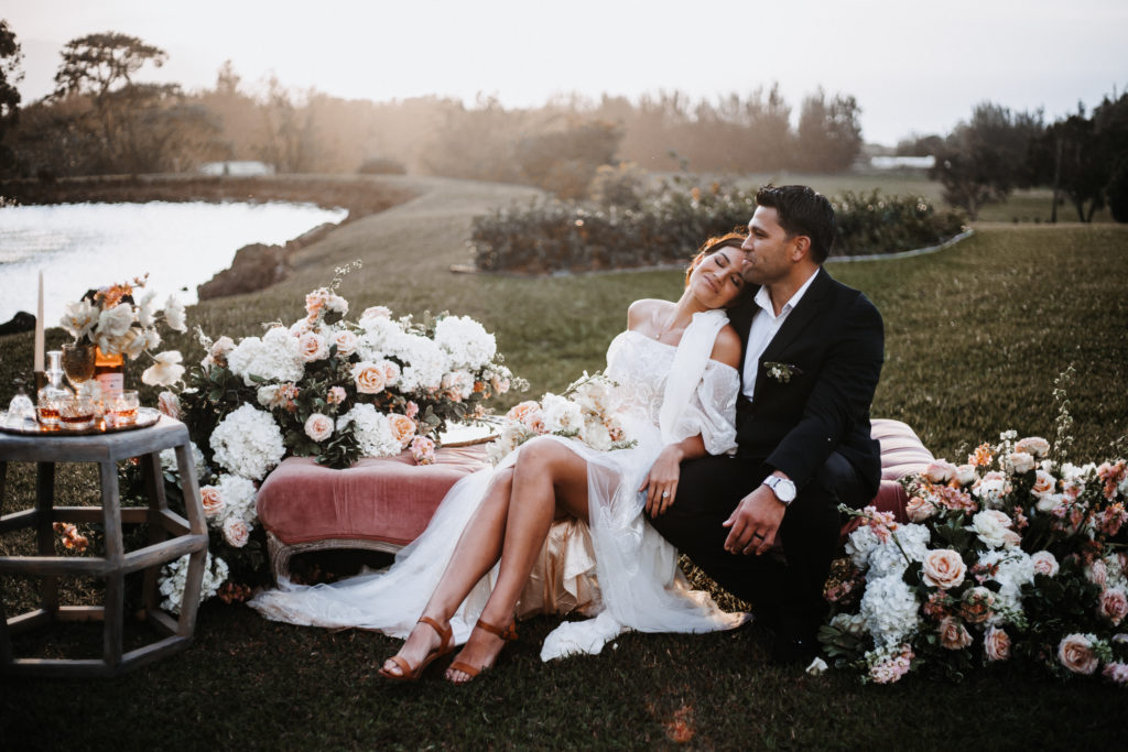 Wedding couple at venue on oahu cuddling on couch with florals in a field