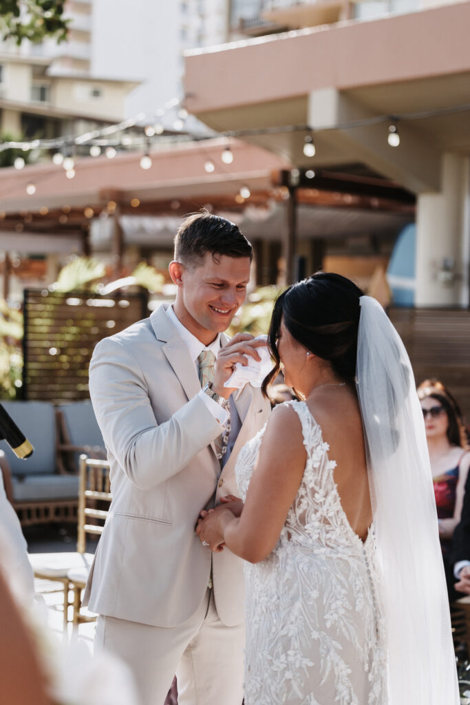 Groom wiping away a tear from bride with the deck waikiki in the background