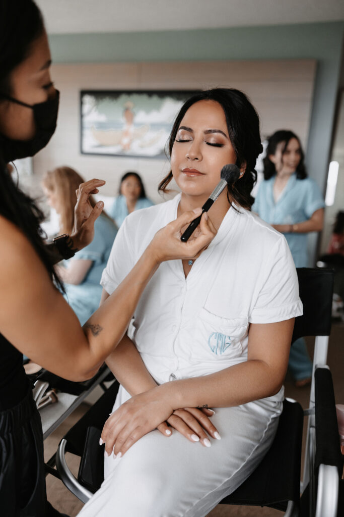 A bride in a white robe has her makeup applied by a professional artist in a room at The Queen Kapiolani Hotel in Waikiki, Honolulu, as her bridal party prepares in the background.