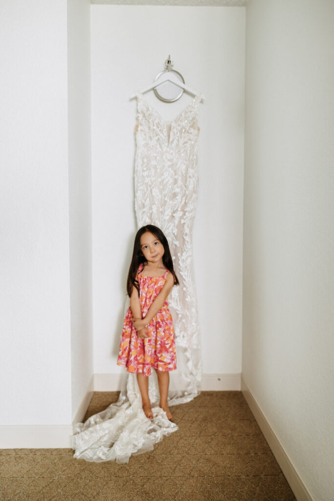 A young girl in a colorful floral dress stands in front of a lace wedding gown hanging on a wall at The Queen Kapiolani Hotel in Waikiki, Honolulu.