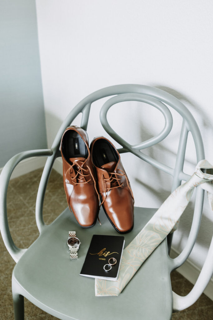 A pair of polished brown leather dress shoes placed on a chair along with a light green tie, a silver watch, wedding rings, and a black card at The Queen Kapiolani Hotel in Waikiki, Honolulu.