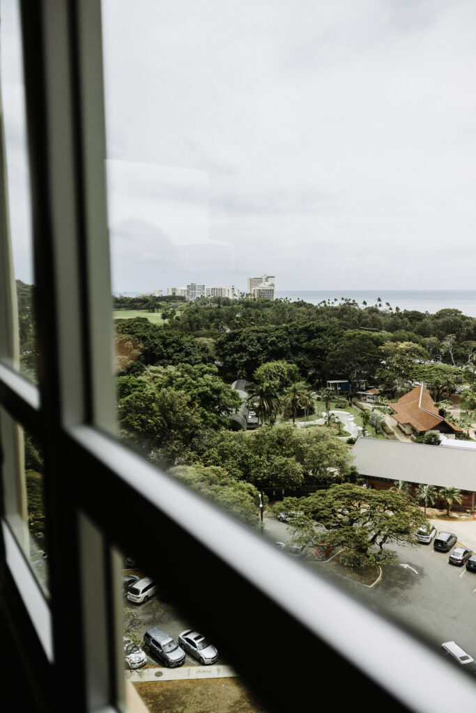 A view through a window at The Queen Kapiolani Hotel in Waikiki, Honolulu, showcasing green parklands, a red-roofed building, and the ocean in the distance.