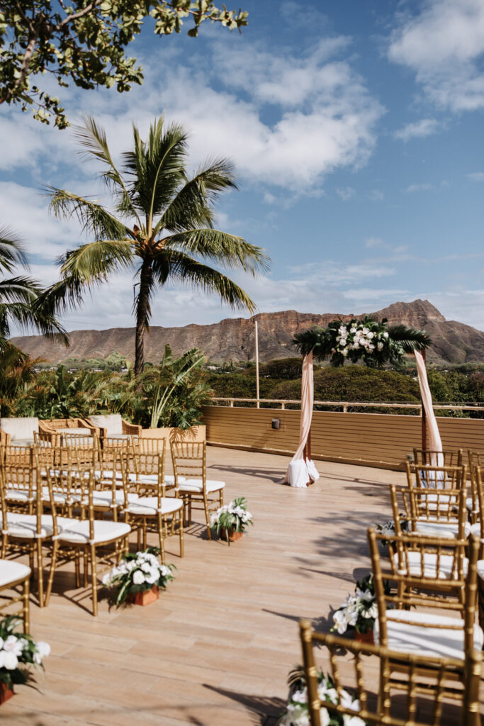 An outdoor wedding ceremony setup with gold chairs, white floral arrangements, and a floral arch, overlooking Diamond Head at The Queen Kapiolani Hotel in Waikiki, Honolulu.