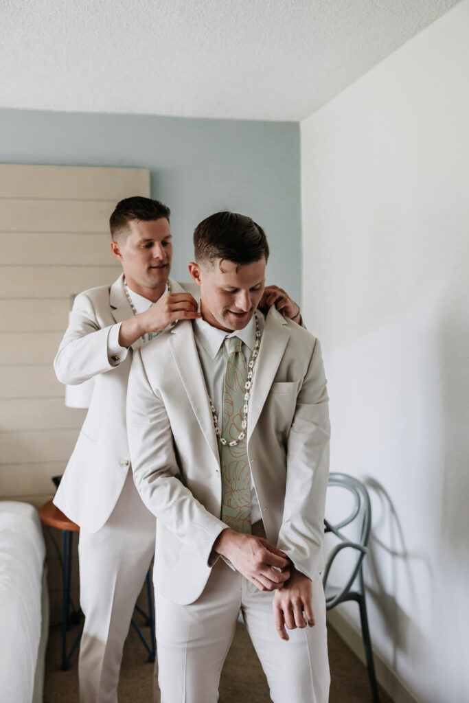 A groom in a cream-colored suit adjusts his sleeve while a groomsman helps place a Hawaiian lei over his shoulders at The Queen Kapiolani Hotel in Waikiki, Honolulu.