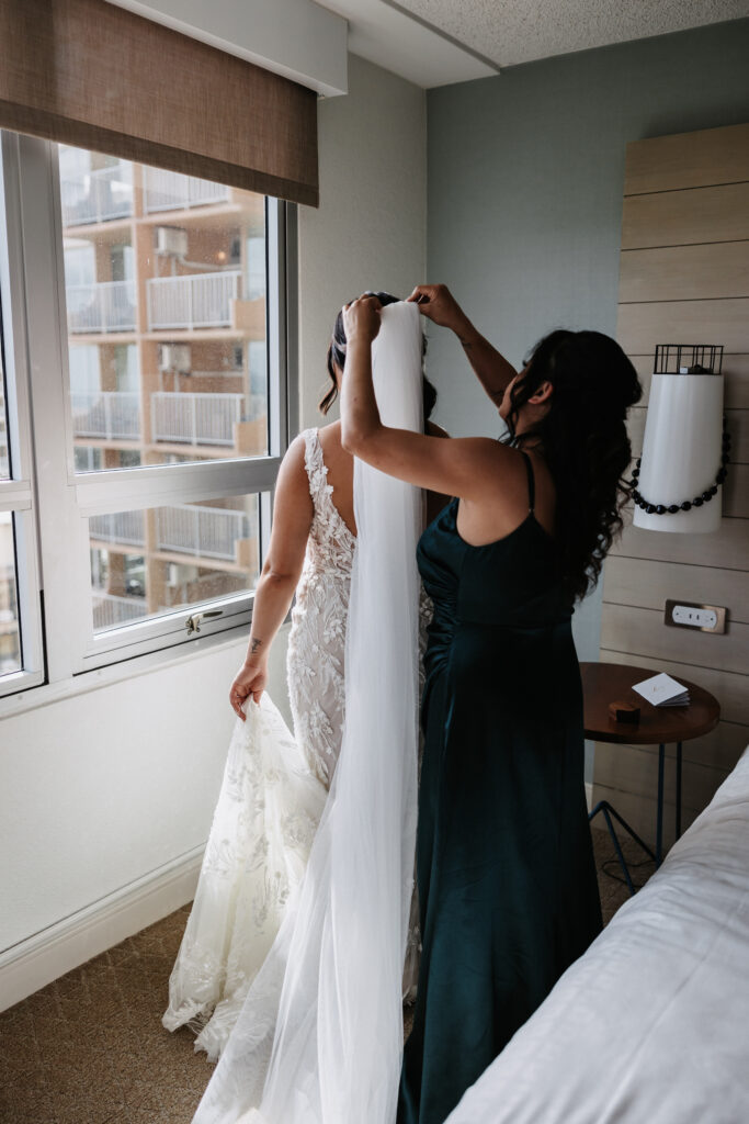 A bride in a lace gown stands near a window while a bridesmaid adjusts her veil at The Queen Kapiolani Hotel in Waikiki, Honolulu.